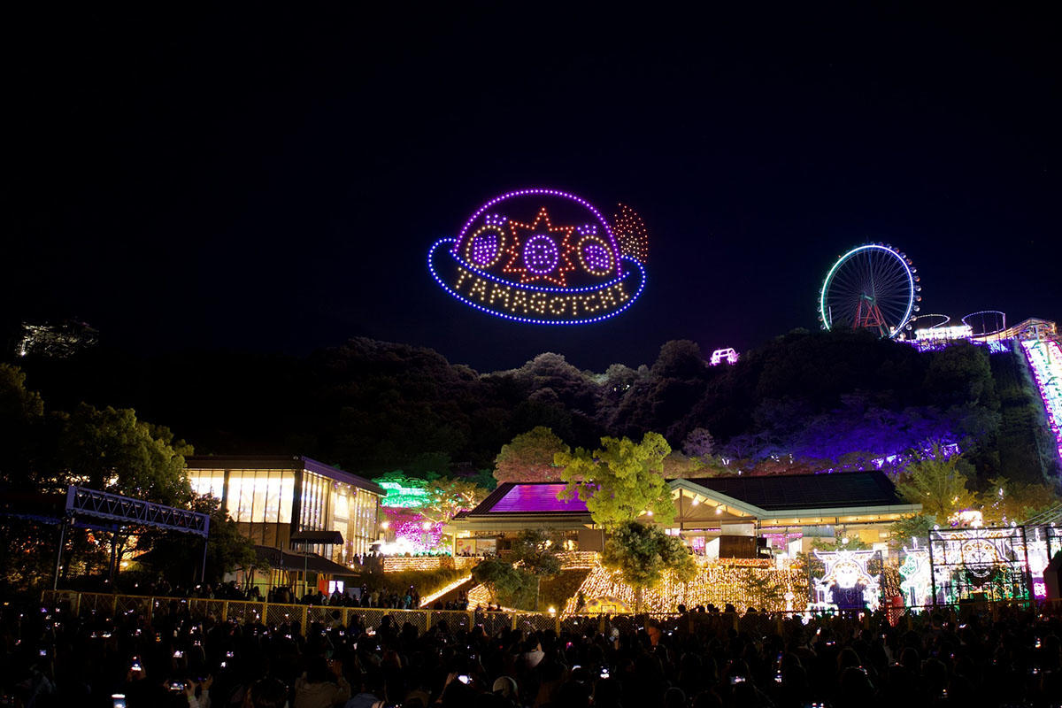 ドローン500機がたまごっちUFOとTAMAGOTCHIのロゴを夜空に描いたシーン