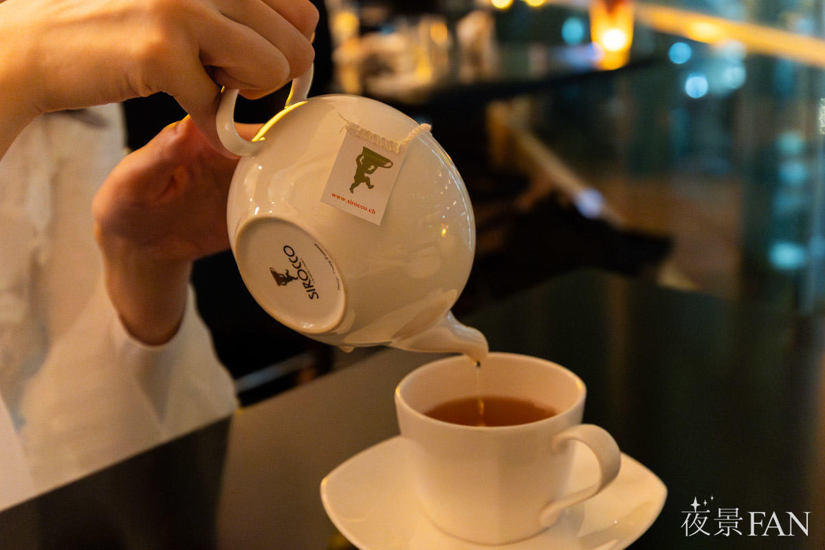 スタッフがSIROCCOのティーポットから紅茶をカップに注ぐ。背景に夜景が広がる