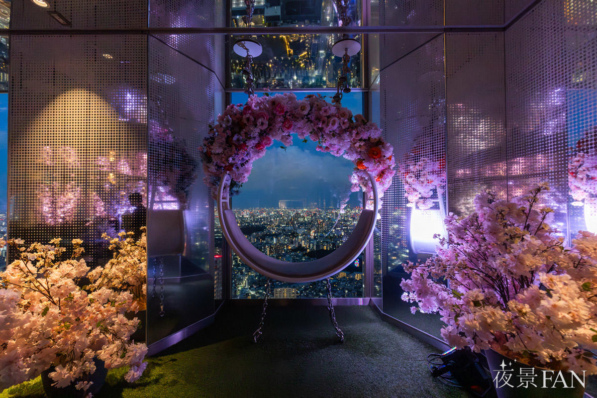 てんぼうパーク さくらまつり2026のブランコ型ベンチ。花のリングと桜の木越しに東京の夜景が広がる